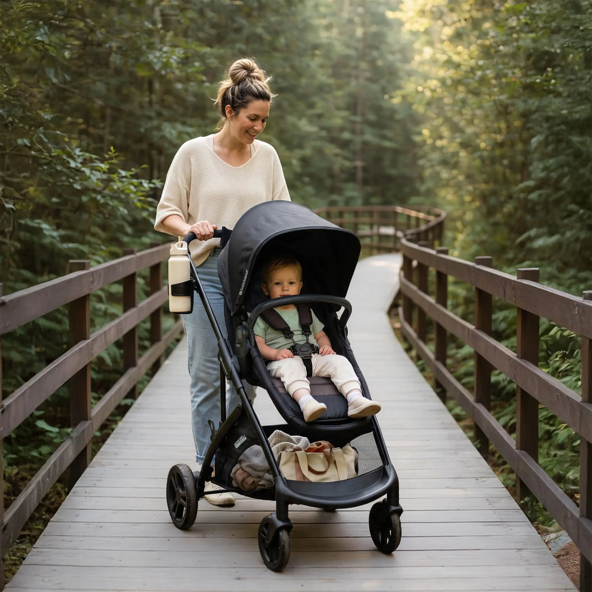 A mom is pushing her child sitting in a Baby Trend Passport Switch 6-in-1 Modular Stroller while going through the woods on a wooden bridge