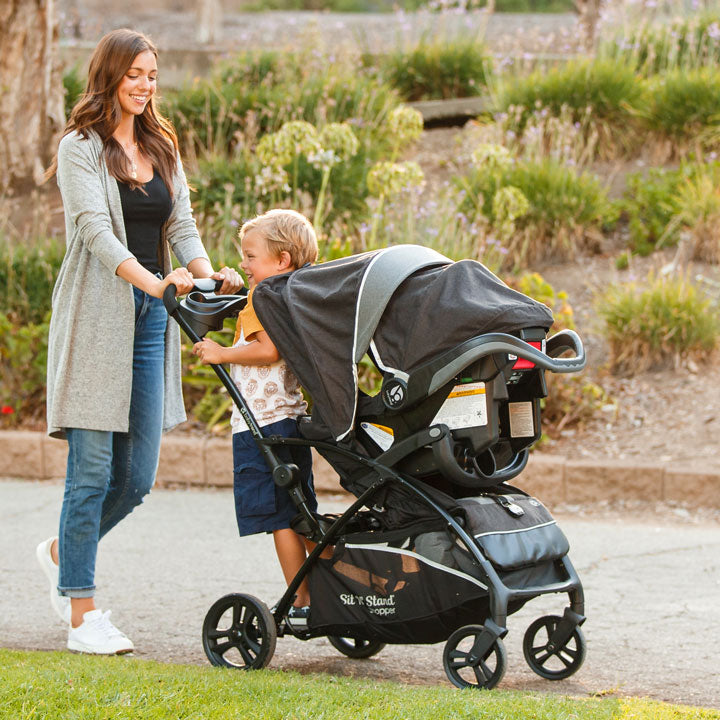 Mom is strolling with her infant in the car seat using the Baby Trend Sit N' Stand Shopper while a toddler stand in the back of the stroller