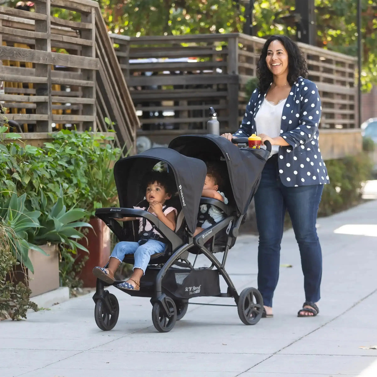 Parent pushing two young children in a Baby Trend Sit N’ Stand Double 2.0 stroller on a sidewalk