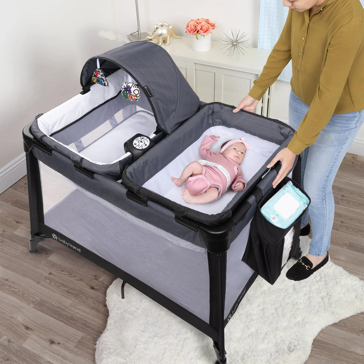 A mom is watching her child laying on the changing table of the Baby Trend Willow Nursery Center Playard inside their living room
