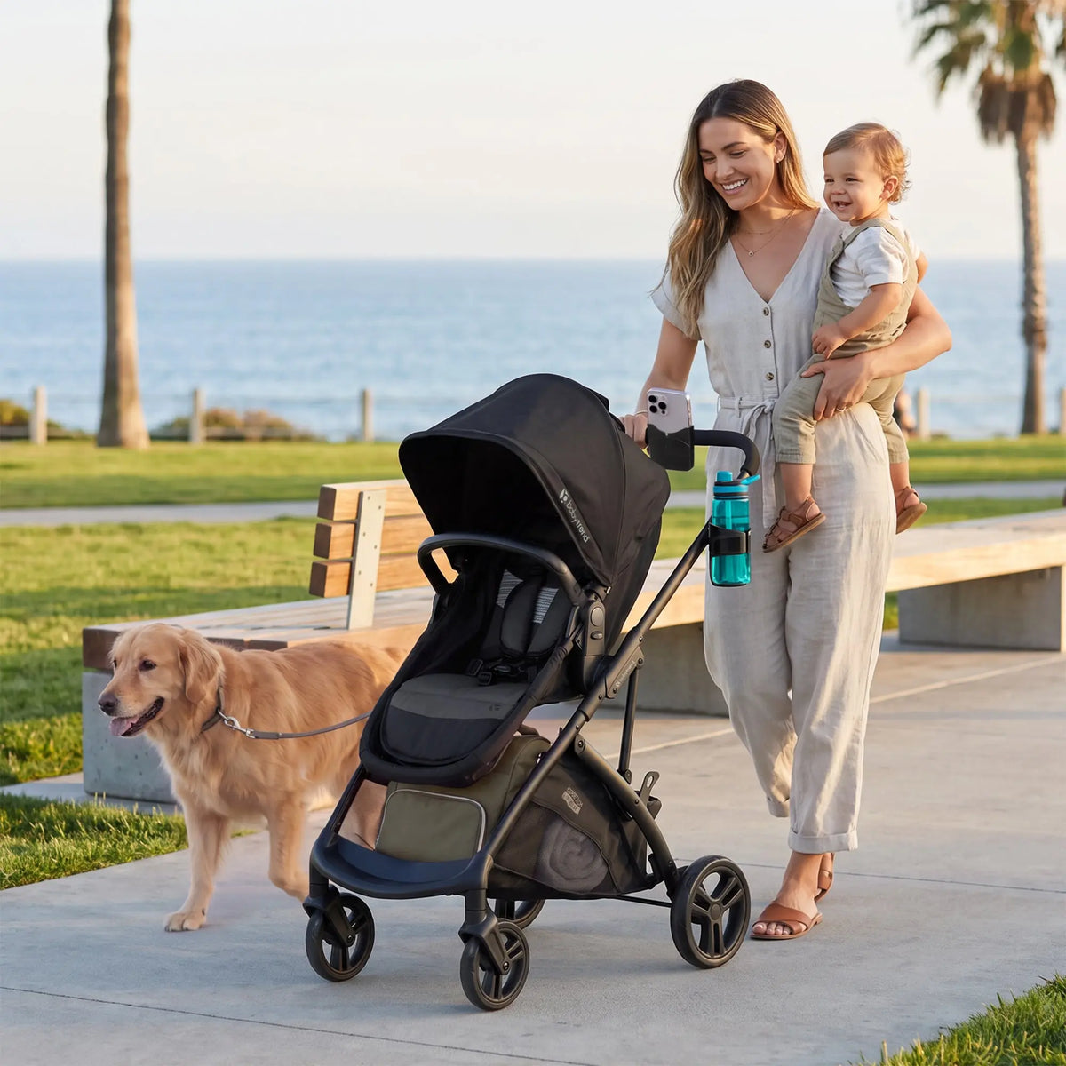 A mother is pushing the Baby Trend Passport Switch 6-in-1 Modular Stroller while carrying her child in the left arm and with their pet dog walking along aside at the beach