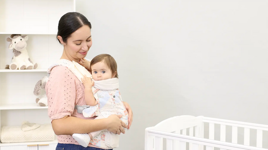 Parent holding a baby in a soft carrier beside a crib